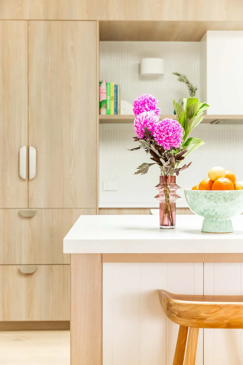 Pink chrysanthemums and a bowl of oranges on a kitchen bench with timber cabinetry