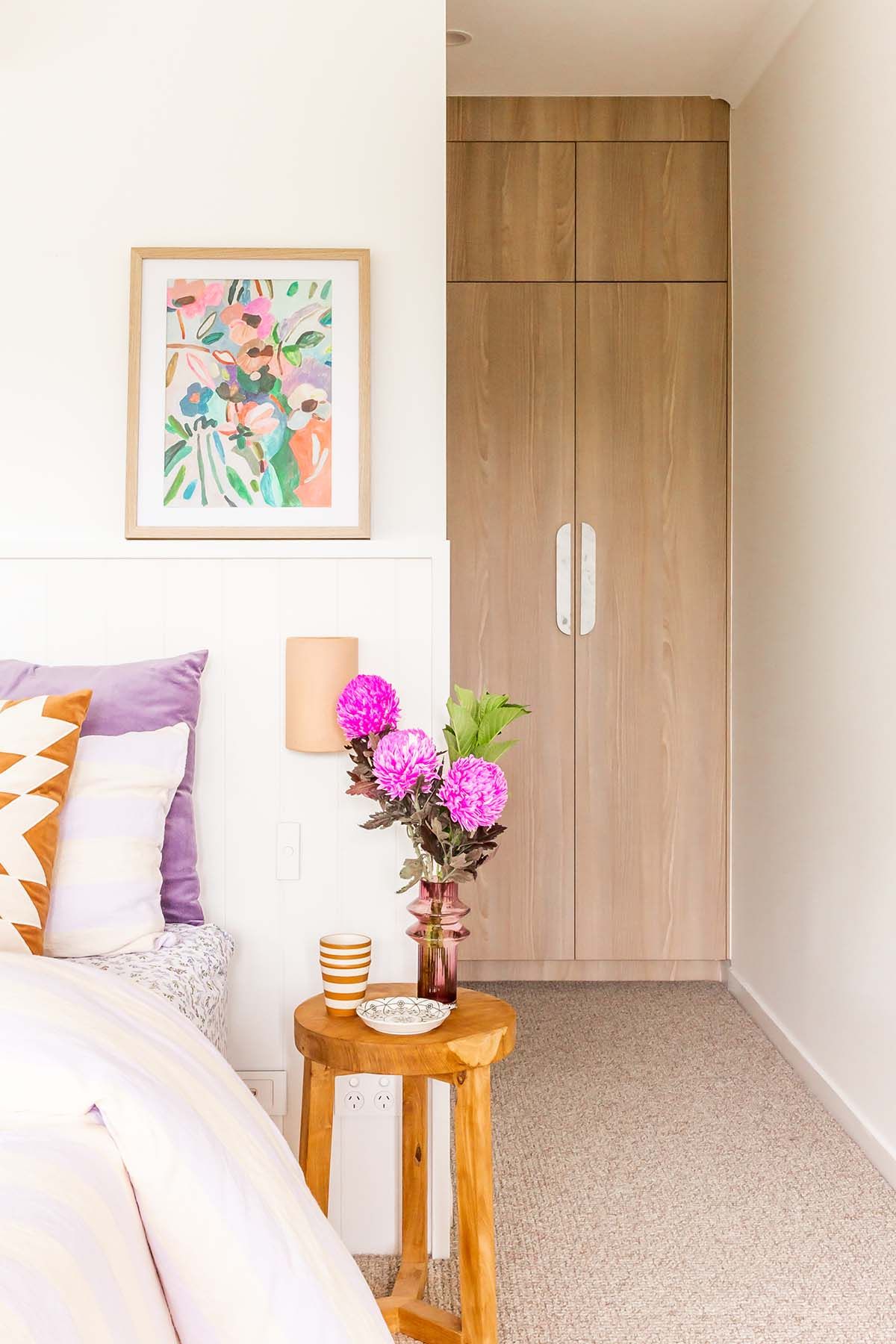 Master bedroom with lilac and patterned cushions, a floral artwork and pink flowers on a timber side table