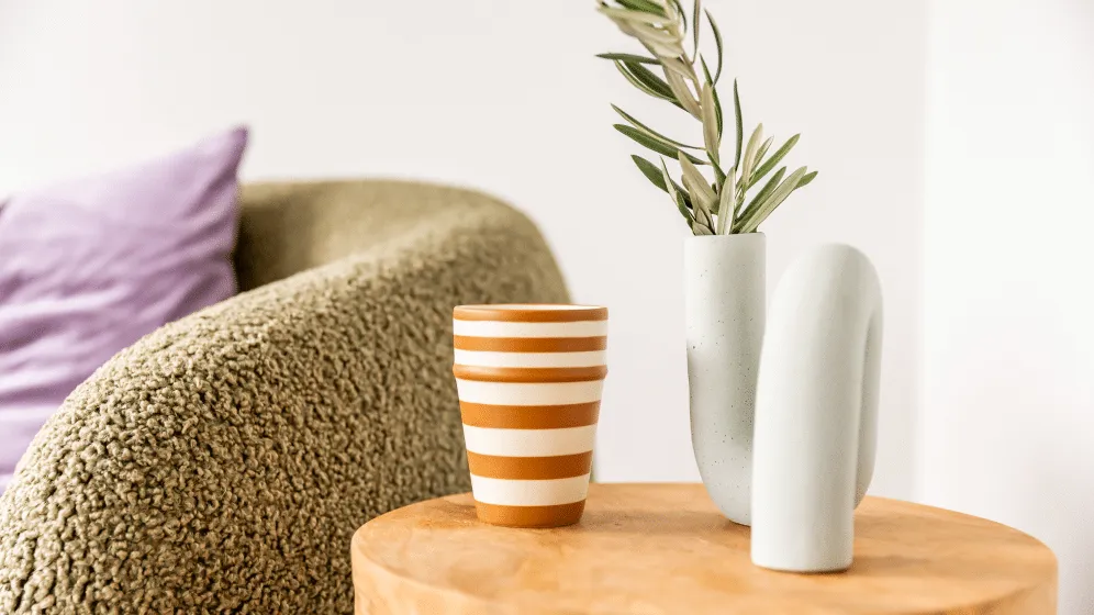 Bouclé armchair with a lilac cushion beside a timber side table with a striped cup and sculptural white vase