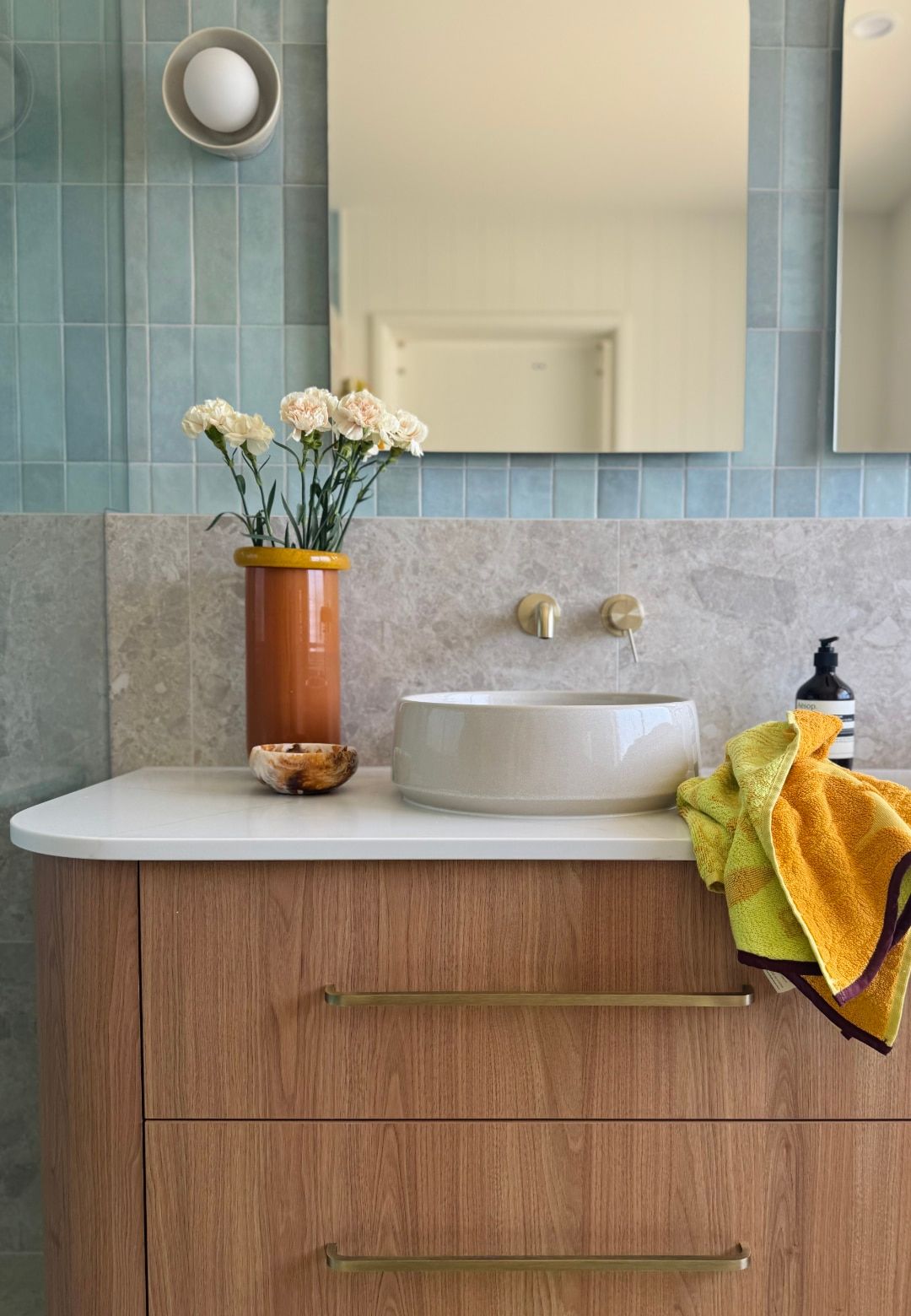Warm timber vanity with a stone basin, brass tapware, fresh flowers and a yellow towel